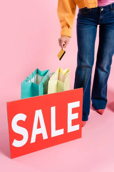 Vista cortada de mulher segurando cartão de crédito perto de sacos de compras e cartão com letras de venda em fundo rosa — Fotografia de Stock