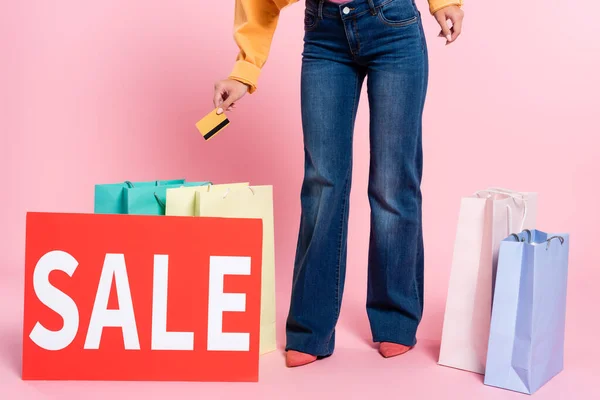 Vista recortada de mujer joven sosteniendo tarjeta de crédito cerca de coloridas bolsas de compras y tarjeta con palabra de venta sobre fondo rosa - foto de stock