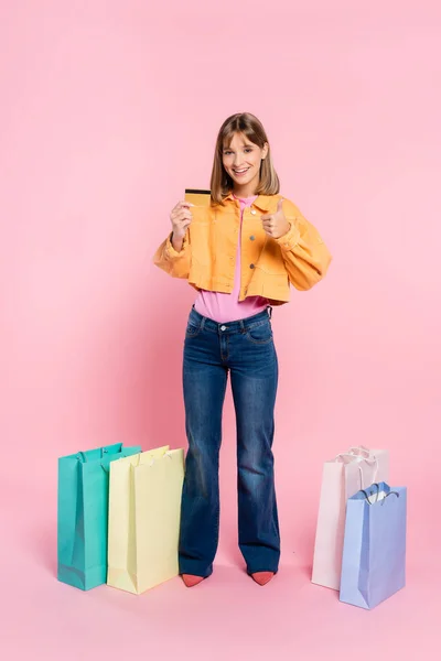 Mulher apontando com o dedo para o cartão de crédito perto de sacos de compras no fundo rosa — Fotografia de Stock