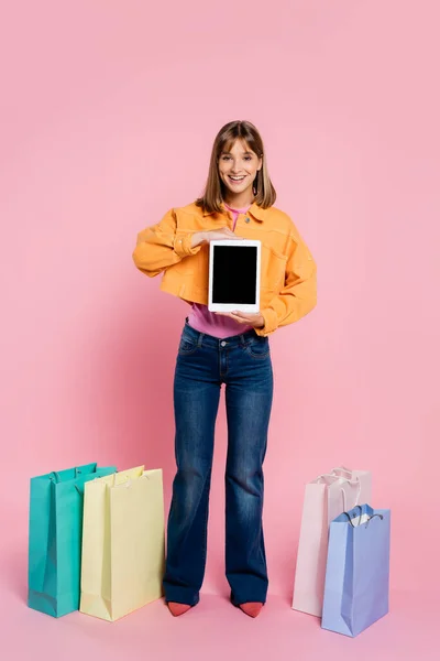 Молодая женщина держит цифровой планшет с чистым экраном возле покупок на розовом фоне — стоковое фото