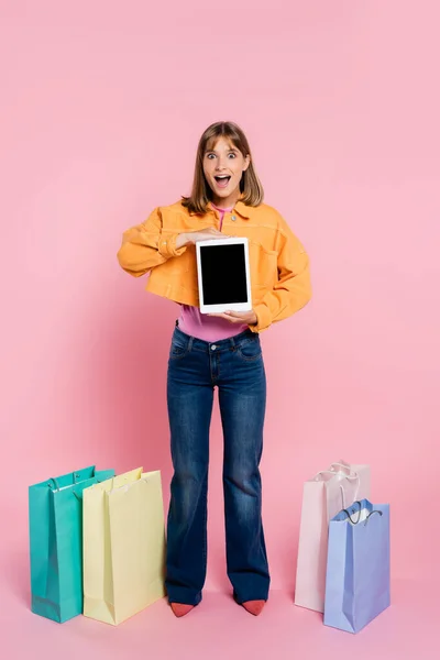 Mujer excitada en chaqueta amarilla sosteniendo tableta digital cerca de coloridas bolsas de compras sobre fondo rosa - foto de stock