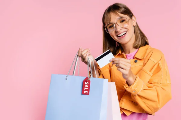 Mujer joven en gafas de sol con tarjeta de crédito y bolsas de compras con letras de venta en la etiqueta de precio sobre fondo rosa - foto de stock