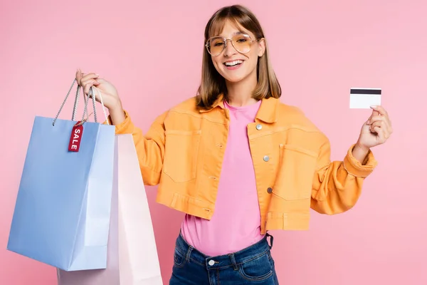 Woman in sunglasses holding shopping bags with sale lettering on price tag and credit card on pink background — Stock Photo