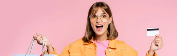 Aufgeregte junge Frau in Sonnenbrille mit Kreditkarte und Einkaufstaschen in rosa — Stockfoto
