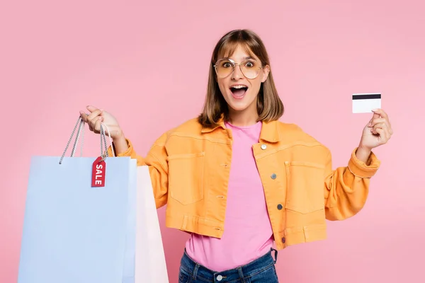 Aufgeregte Frau in Sonnenbrille mit Kreditkarte und Einkaufstaschen mit Verkaufswort auf Preisschild auf rosa Hintergrund — Stockfoto