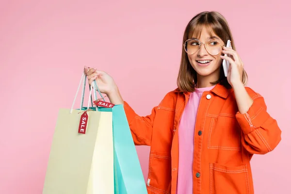 Mujer joven hablando en el teléfono inteligente mientras sostiene bolsas de compras con etiquetas de precios aislados en rosa - foto de stock