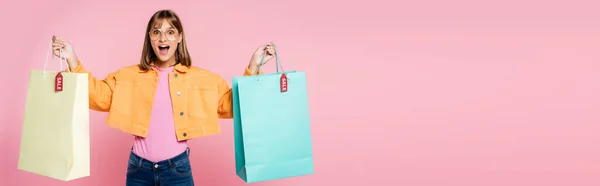 Cultivo panorámico de mujer conmocionada sosteniendo bolsas de compras con etiquetas de precios sobre fondo rosa - foto de stock