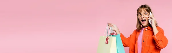 Panoramic shot of surprised woman holding shopping bags with price tags while talking on smartphone on pink background — Stock Photo