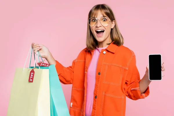 Mujer emocionada mostrando teléfono inteligente con pantalla en blanco mientras sostiene bolsas de compras con etiquetas de precios aislados en rosa - foto de stock