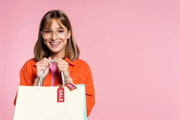 Jeune femme en lunettes de soleil regardant la caméra et tenant des sacs à provisions avec mot de vente sur les étiquettes de prix isolé sur rose — Photo de stock