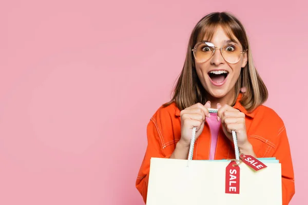 Mulher excitada em óculos de sol segurando sacos de compras com palavra venda em etiquetas de preço isolado em rosa — Fotografia de Stock
