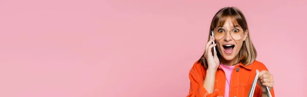 Панорамний знімок здивованої жінки в сонцезахисних окулярах дивиться на камеру, розмовляючи на смартфоні ізольовано на рожевому — стокове фото