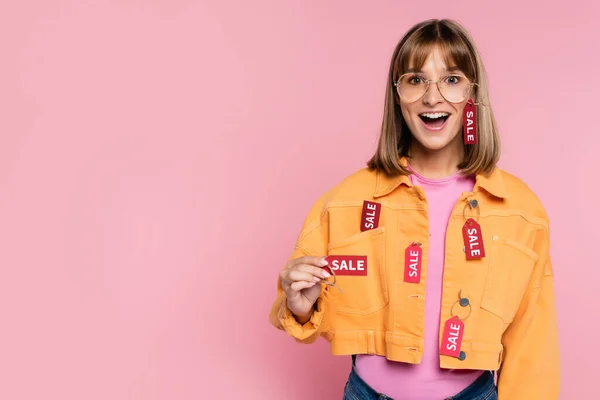 Mujer emocionada mostrando etiqueta de precio con palabra venta en la cámara sobre fondo rosa - foto de stock