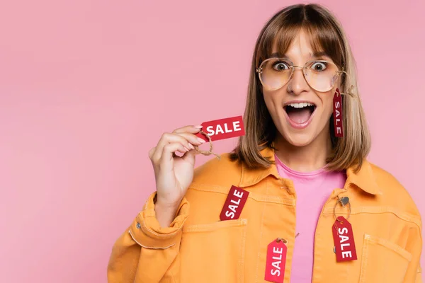 Schockierte Frau mit Sonnenbrille und gelber Jacke hält Preisschild mit Verkaufswort auf rosa — Stockfoto