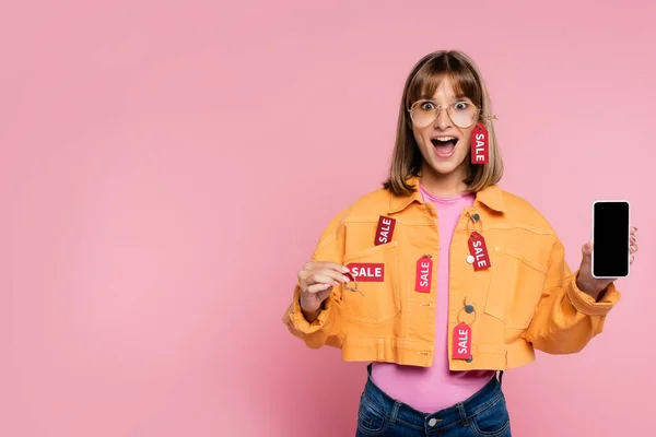 Mujer sorprendida sosteniendo teléfono inteligente con pantalla en blanco y etiqueta de precio con letras de venta sobre fondo rosa - foto de stock