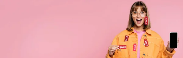 Изображение шокированной женщины с ценником и смартфоном на розовом фоне — стоковое фото