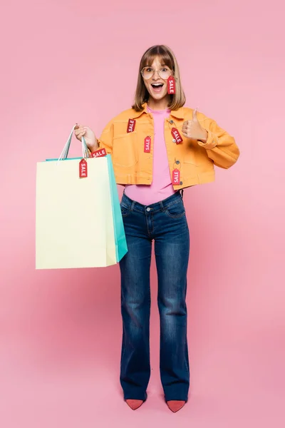 Aufgeregte Frau hält Einkaufstüten mit Preisschildern in der Hand und zeigt Daumen nach oben auf rosa Hintergrund — Stockfoto