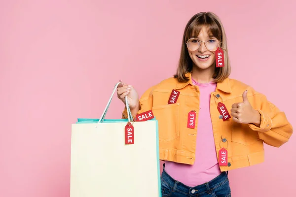 Mulher com etiquetas de preço no casaco segurando sacos de compras e mostrando como gesto isolado em rosa — Fotografia de Stock