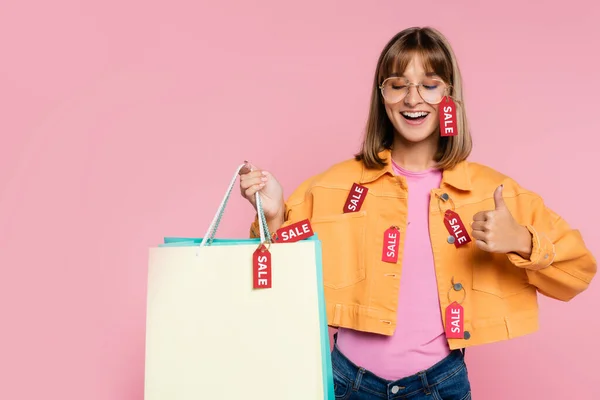 Mulher com olhos fechados segurando sacos de compras com etiquetas de preço e mostrando polegar para cima isolado em rosa — Fotografia de Stock