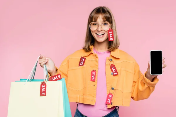 Mujer joven sosteniendo bolsas de compras con etiquetas de precio y venta de letras y teléfono inteligente con pantalla en blanco sobre fondo rosa - foto de stock