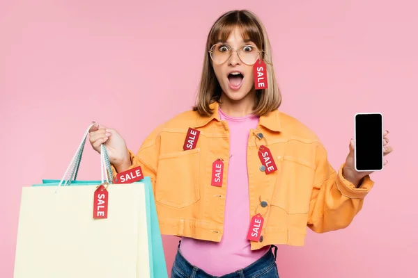 Femme choquée dans les lunettes de soleil et les étiquettes de prix sur la veste montrant smartphone et sacs à provisions colorés sur fond rose — Photo de stock