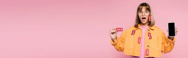 Colpo panoramico di donna scioccata in occhiali da sole che mostra il cartellino del prezzo con parola di vendita e smartphone con schermo bianco su sfondo rosa — Stock Photo