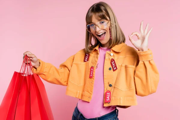 Жінка з ціновими тегами на куртці, що відкриває червоні сумки для покупок і показує нормальний жест на рожевому фоні — стокове фото