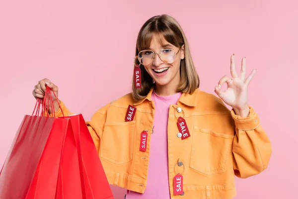 Mulher de jaqueta com etiquetas de preço segurando sacos de compras e mostrando gesto ok no fundo rosa — Fotografia de Stock