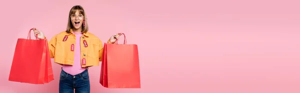 Imagem horizontal da mulher excitada com etiquetas de preço na jaqueta e óculos de sol olhando para a câmera enquanto segurava sacos de compras vermelhos no fundo rosa — Fotografia de Stock