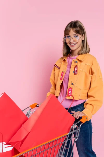 Junge Frau in Jacke mit Verkaufsbuchstaben auf Preisschildern blickt in die Kamera in der Nähe von Einkaufswagen mit Einkaufstaschen auf rosa Hintergrund — Stockfoto