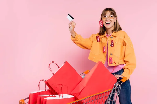 Захоплена жінка з ціновими тегами на куртці тримає кредитну картку біля сумки в кошику на рожевому фоні — стокове фото