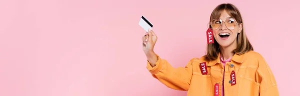 Panoramic shot of excited woman in jacket with price tags holding credit card on pink background — Stock Photo