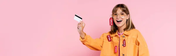 Cosecha horizontal de mujer joven excitada en gafas de sol y chaqueta con etiquetas de precios con tarjeta de crédito sobre fondo rosa - foto de stock