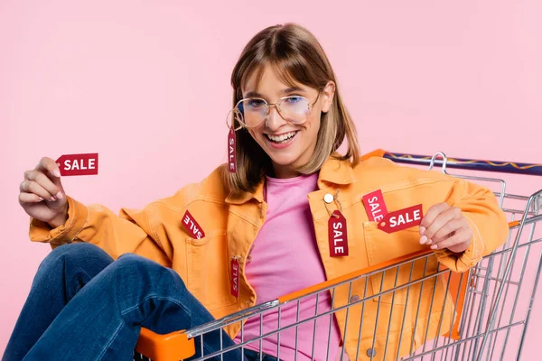 Junge Frau zeigt Preisschilder mit Verkaufsbuchstaben, während sie im Einkaufswagen isoliert auf pink sitzt — Stockfoto