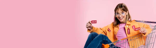 Panoramic concept of woman showing price tags with sale word while sitting in cart on pink background — Stock Photo