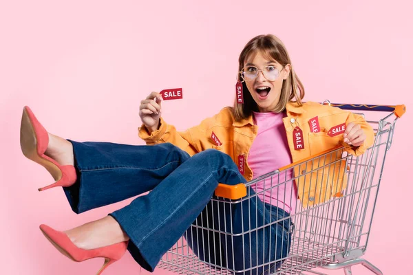 Enfoque selectivo de mujer impactada sosteniendo etiquetas de precios con letras de venta mientras está sentada en el carrito de la compra sobre fondo rosa - foto de stock