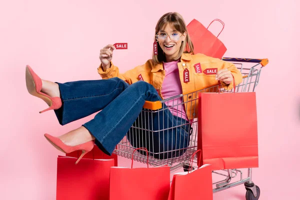 Young woman holding price tag with sale lettering and red shopping bag while sitting in cart on pink background — Stock Photo