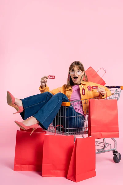 Femme choquée en talons tenant des étiquettes de prix tout en étant assis dans le panier près de sacs à provisions sur fond rose — Photo de stock