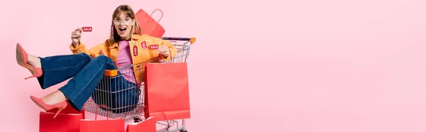 Horizontale Ernte überraschter Frau mit Preisschildern, die in den Einkaufswagen mit Einkaufstaschen auf rosa Hintergrund blickt — Stockfoto