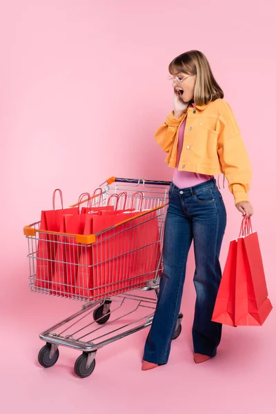 Mujer sorprendida con la mano cerca de la mejilla mirando las bolsas de la compra en el carrito sobre fondo rosa - foto de stock