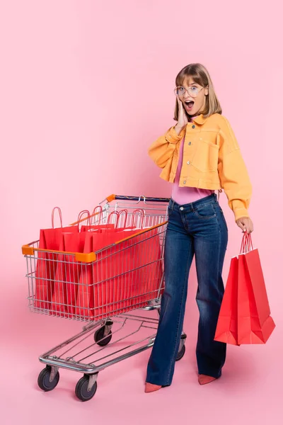 Donna scioccata in piedi vicino al carrello con borse della spesa rosse su sfondo rosa — Foto stock