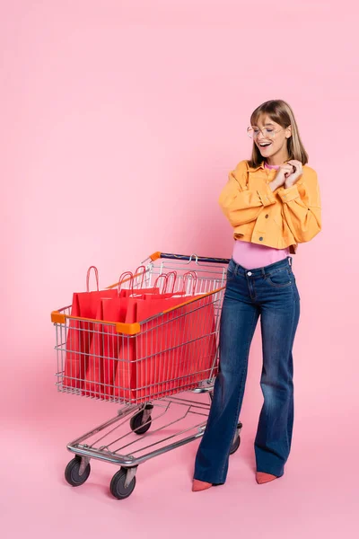 Jovem mulher em óculos de sol olhando para sacos de compras vermelhos no carrinho no fundo rosa — Fotografia de Stock