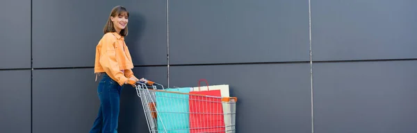 Conceito panorâmico de jovem olhando para a câmera perto de sacos de compras no carrinho e construção — Fotografia de Stock