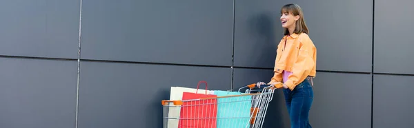 Conceito panorâmico de mulher excitada andando perto do carrinho com sacos de compras e fachada do edifício — Fotografia de Stock