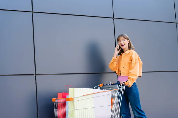 Mujer hablando por celular cerca de bolsas de compras en carro y fachada de edificio - foto de stock