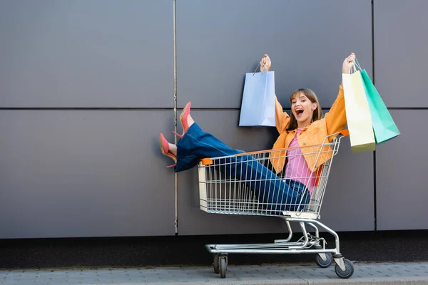 Excited woman in heels holding colorful shopping bags while sitting in cart near building — Stock Photo