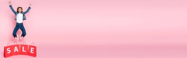 Панорамне зображення збудженої жінки, що стрибає біля сумки з продажем на рожевому фоні — стокове фото