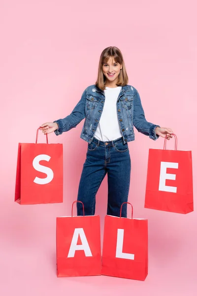 Donna in giacca di denim guardando la fotocamera mentre tiene borse della spesa rosse con scritte in vendita su sfondo rosa — Foto stock