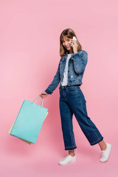 Jovem mulher de jaqueta jeans falando no smartphone e segurando sacos de compras no fundo rosa — Fotografia de Stock