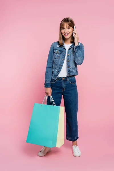 Young woman looking at camera while talking on cellphone and holding shopping bags on pink background — Stock Photo